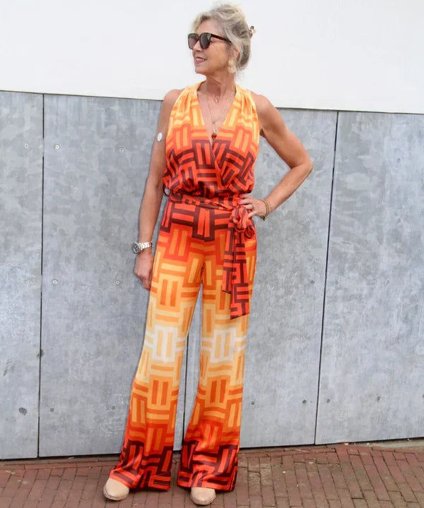 Vrouw draagt een kleurrijke jumpsuit met oranje print, V-hals, tailleceintuur, wijde pijpen, zonnebril en lichte schoenen.